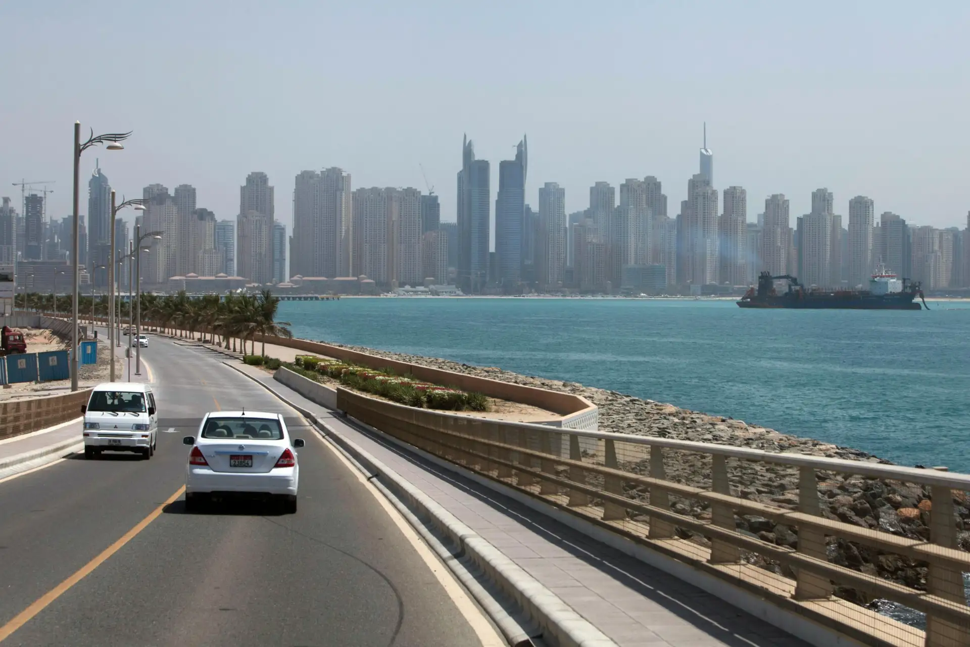 Coastal road with two cars driving, palm trees lining the side, blue sea to the right, and a city skyline with tall buildings in the background.