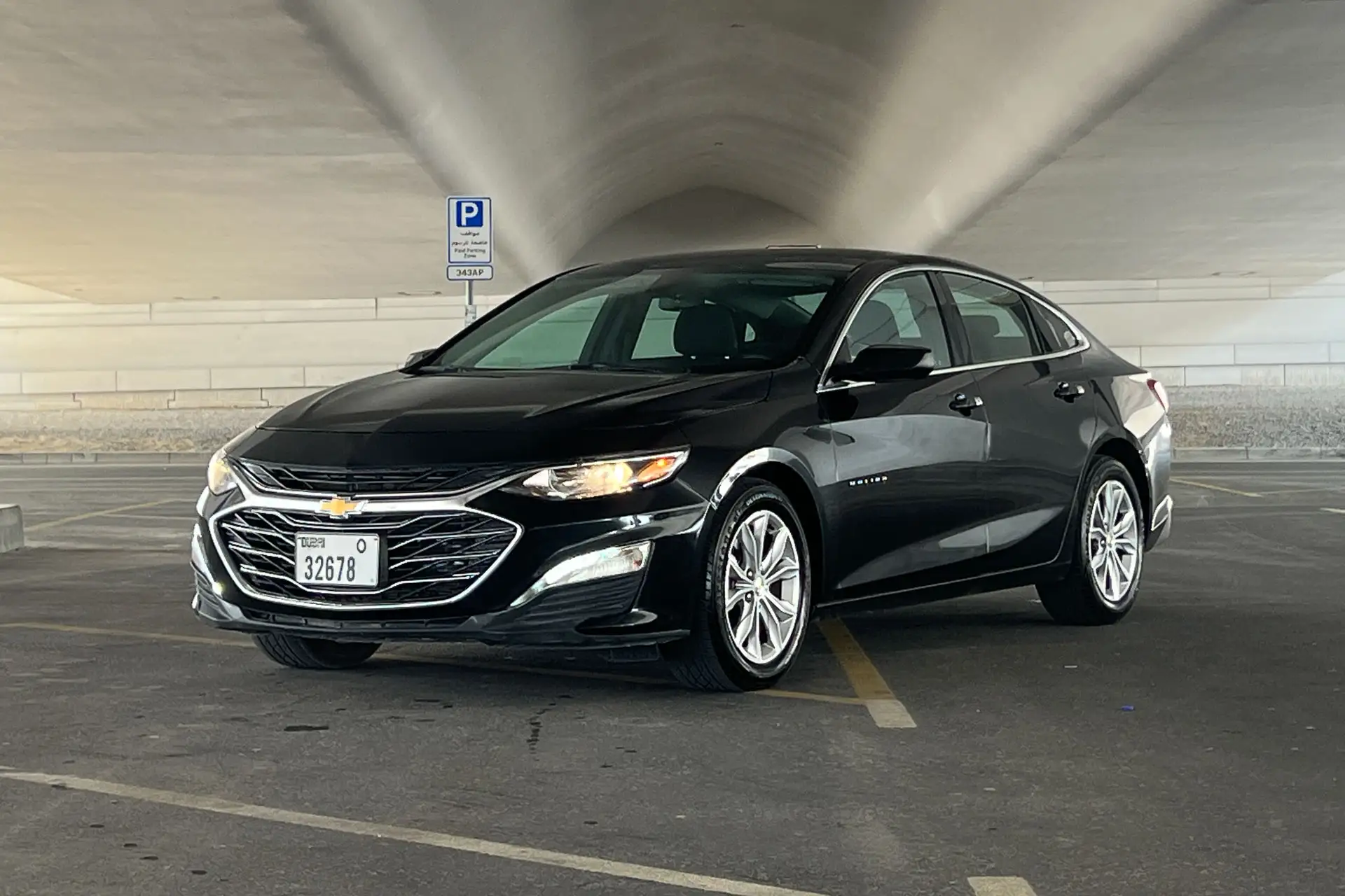 A black Chevrolet sedan parked in an indoor parking lot with a ceiling archway. A "Parking" sign is visible nearby.