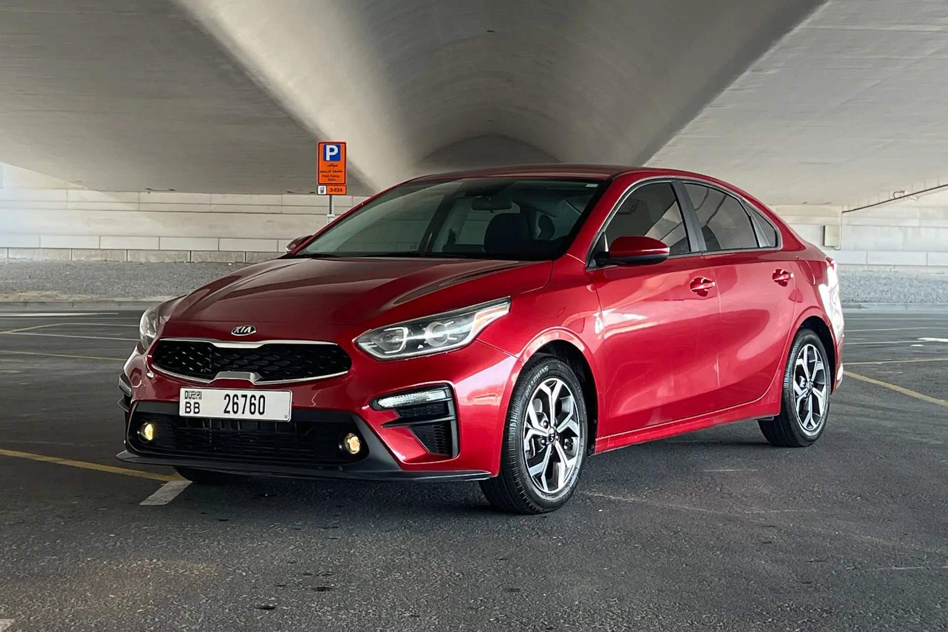 Red Kia sedan parked under a large concrete structure in an empty parking area, with a clear view of license plates and modern headlights.