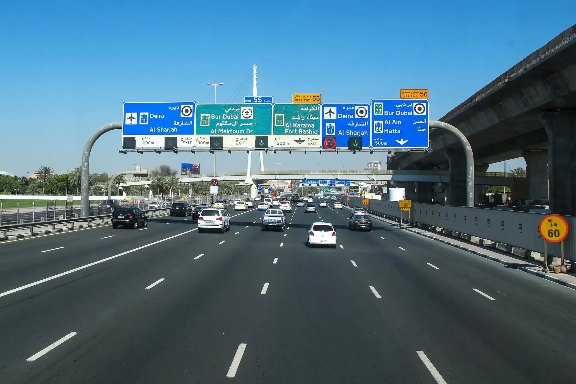 Highway with multiple lanes and road signs in Dubai, directing to Deira, Bur Dubai, Al Sharjah, Al Ain, and Hatta under a clear blue sky.