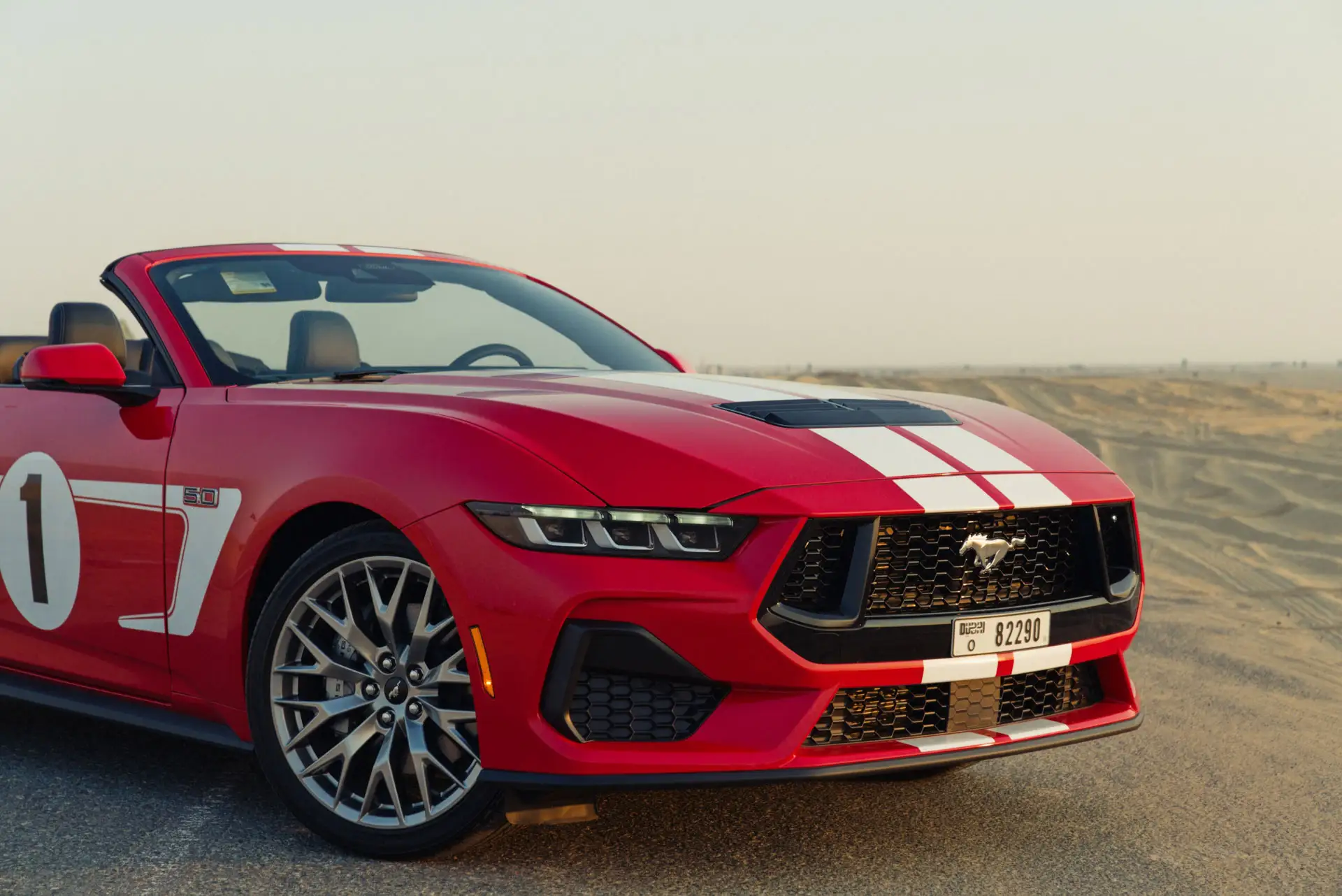 Red convertible sports car with white racing stripes parked on a desert road, featuring a sleek front design and distinctive emblem.