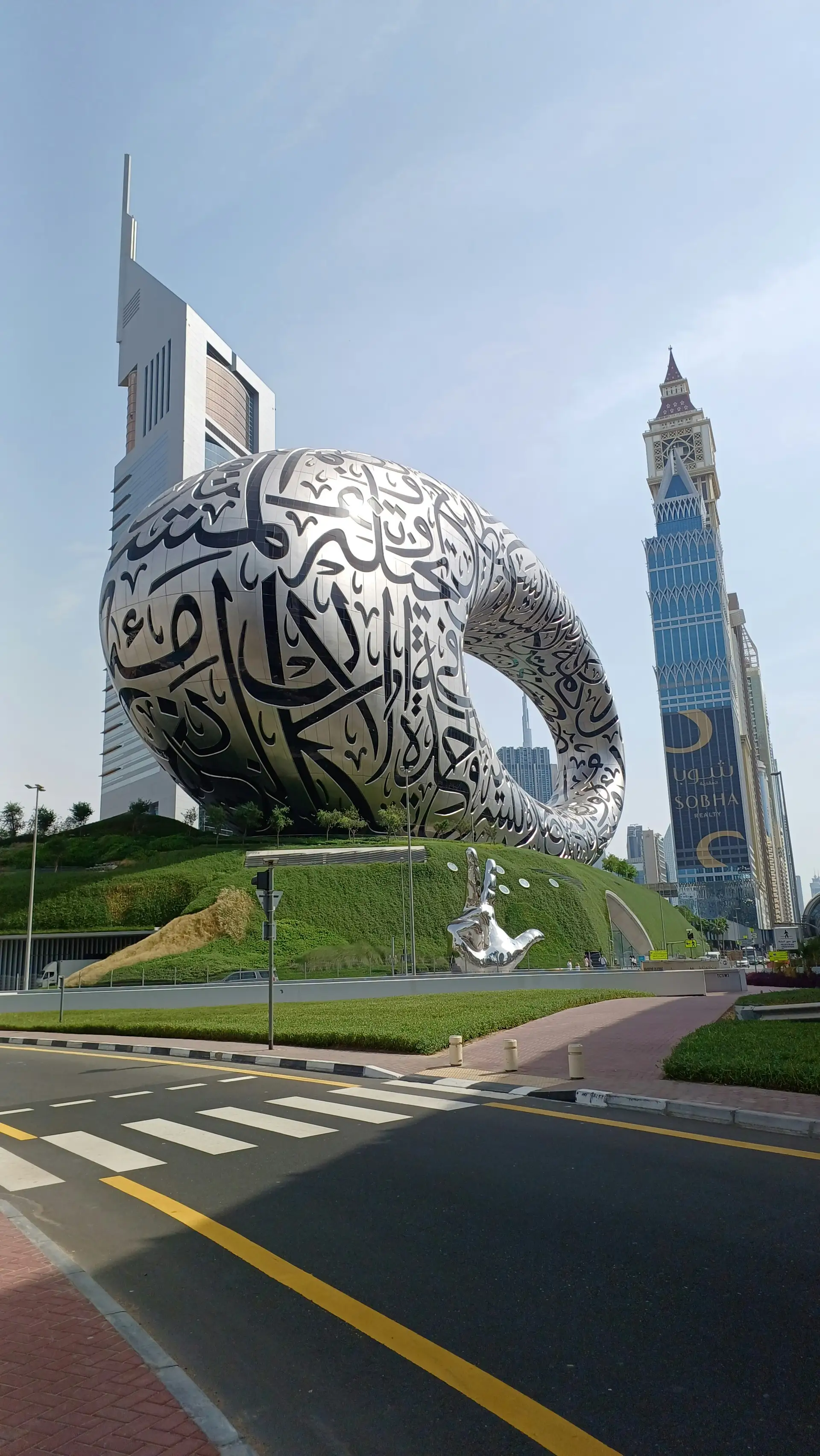 Futuristic building with Arabic calligraphy, shaped like an oval ring, set against a cityscape with modern skyscrapers under a clear sky.