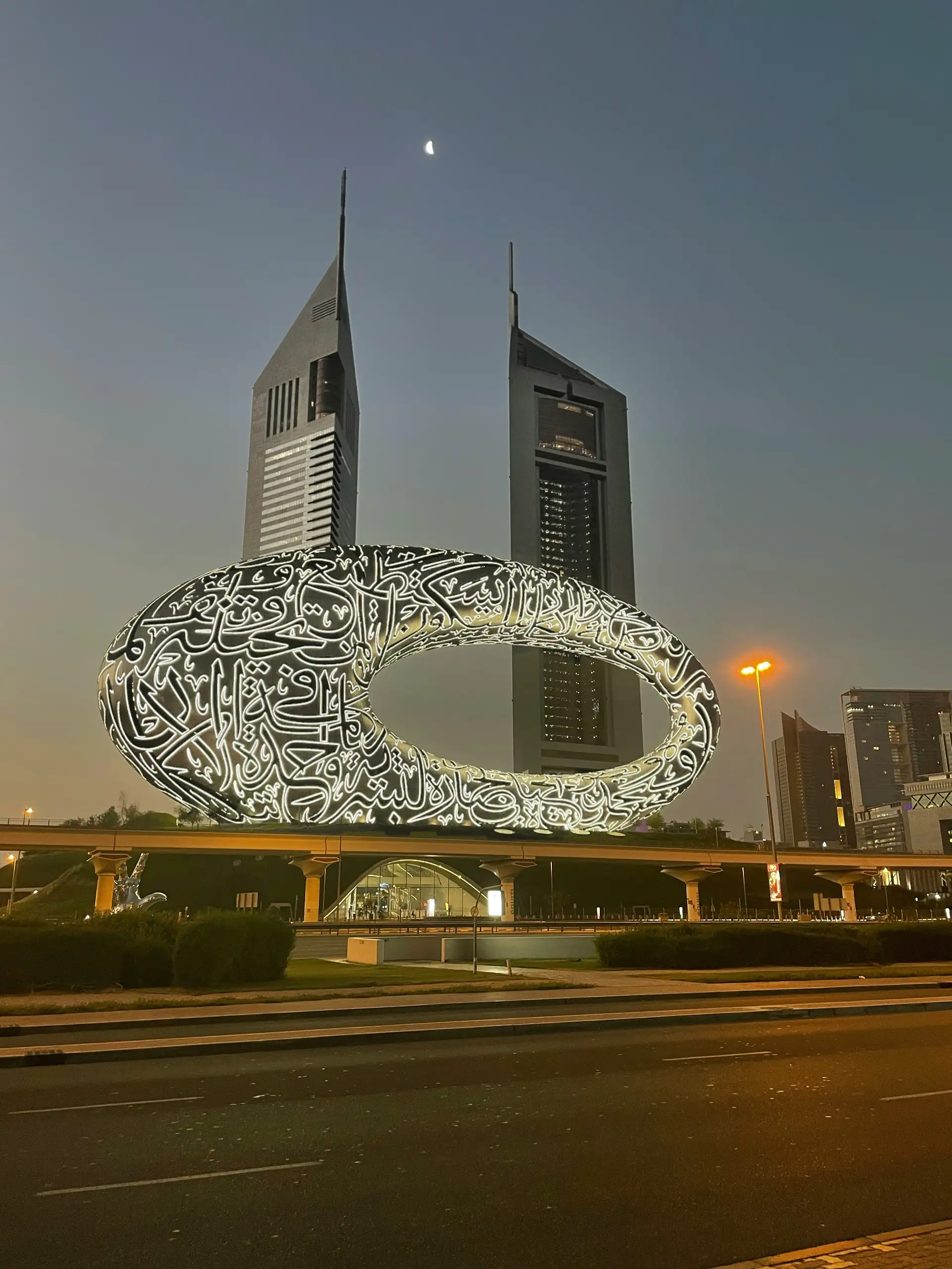 The Museum of the Future in Dubai, with its illuminated Arabic calligraphy, stands in front of two skyscrapers at dusk.