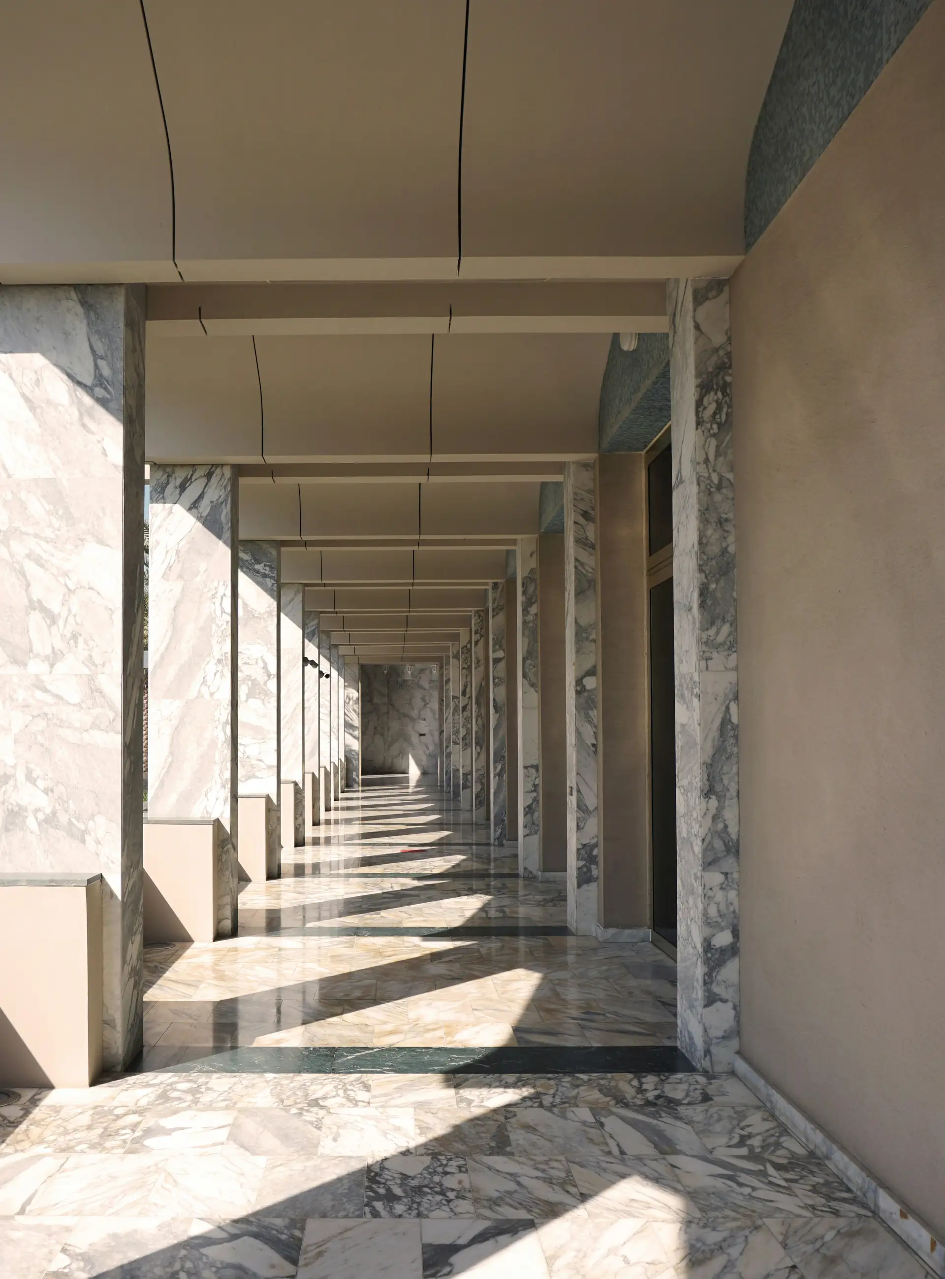 Sunlit corridor with marble columns and floor, featuring long shadows and a vanishing point perspective in a modern architectural setting.