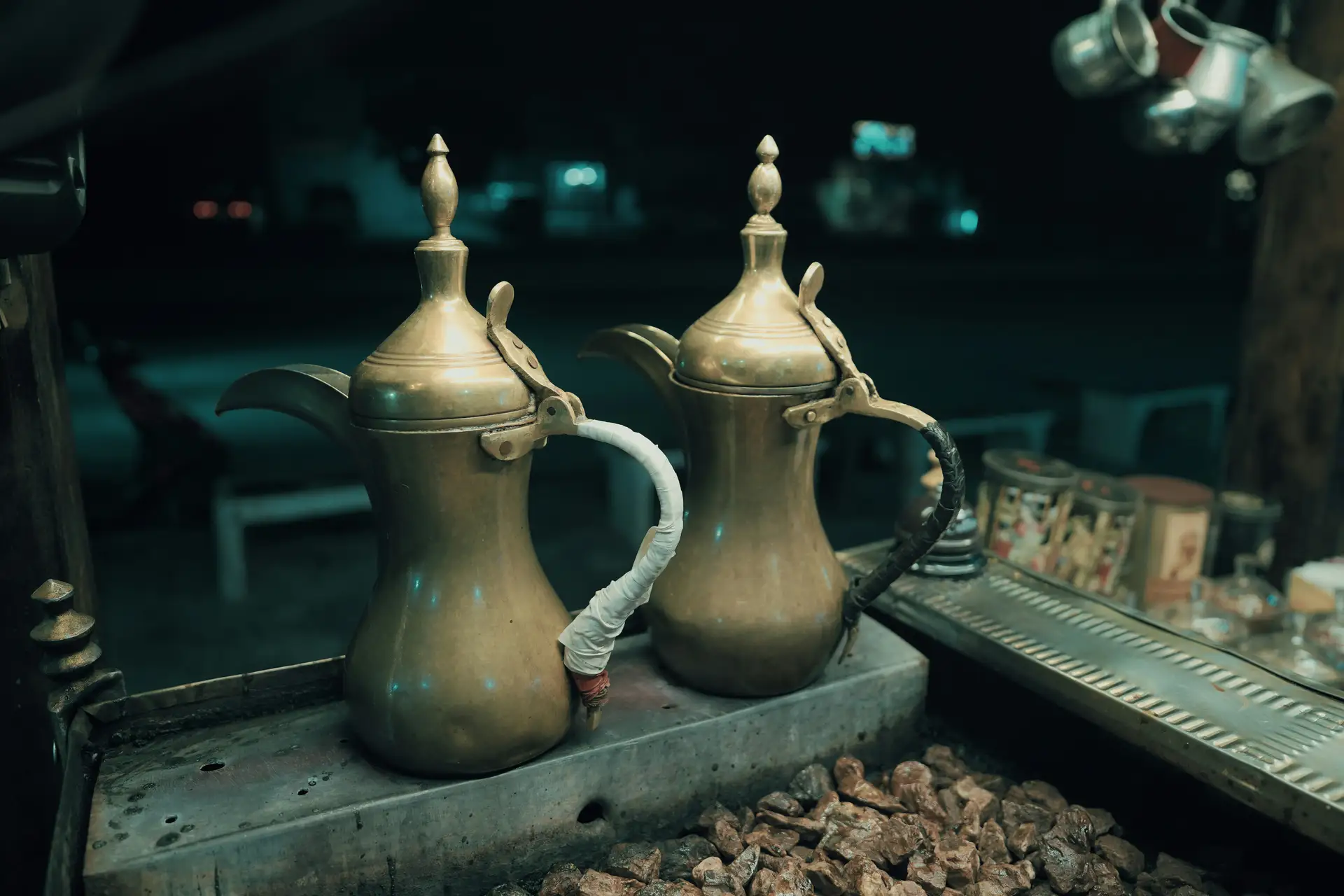 Two ornate brass coffee pots on a bed of hot coals, with a metal counter and hanging cups in the background.