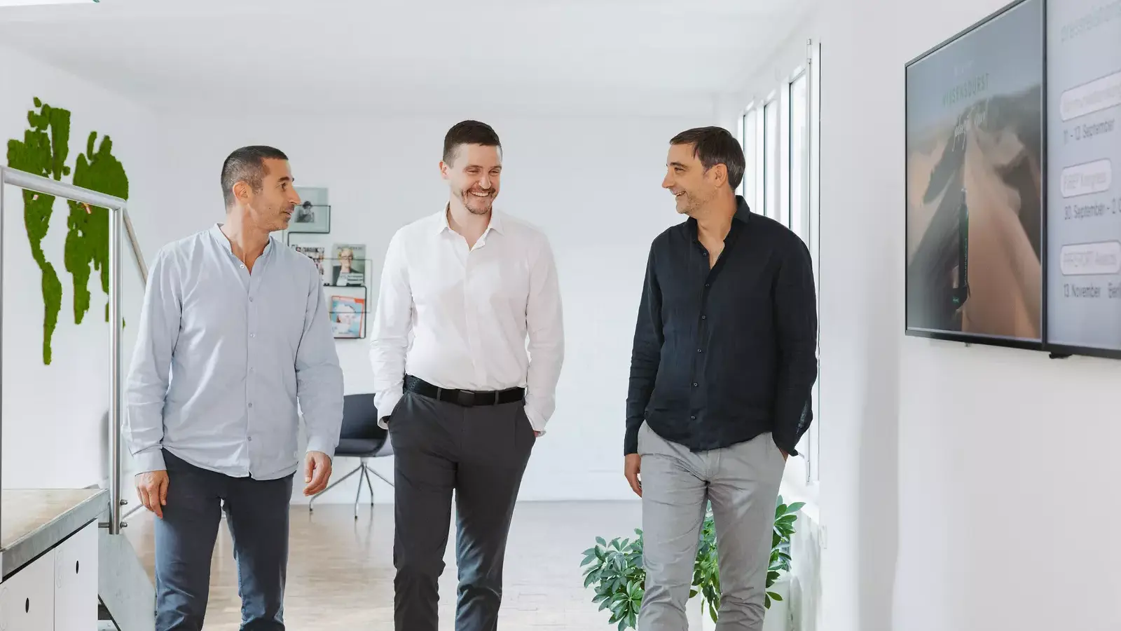Three men in business casual attire walk and converse in a bright office corridor with a plant and a wall-mounted screen displaying information.