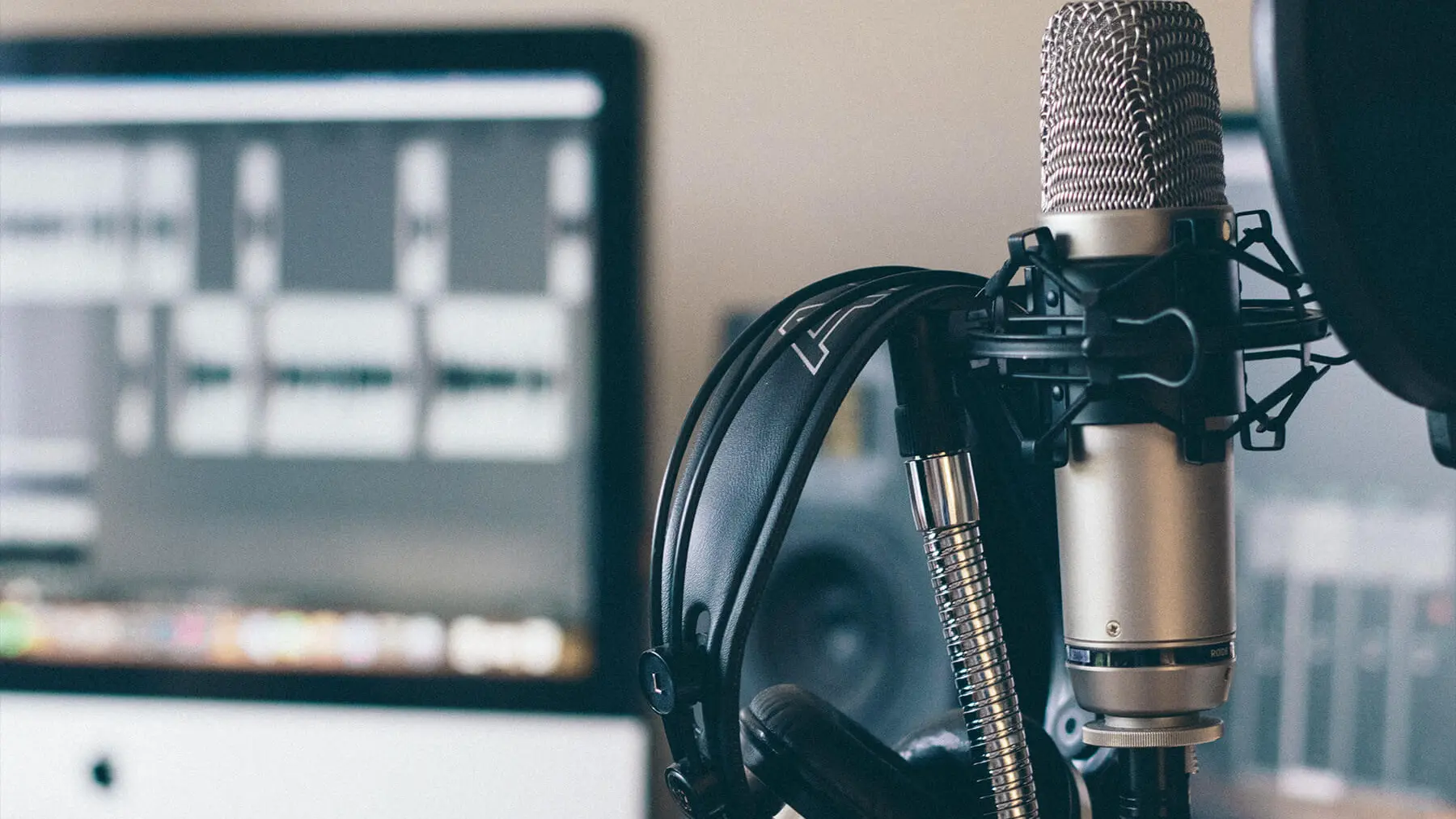 Microphone with attached headphones in focus, placed in front of a blurred computer screen displaying audio editing software.