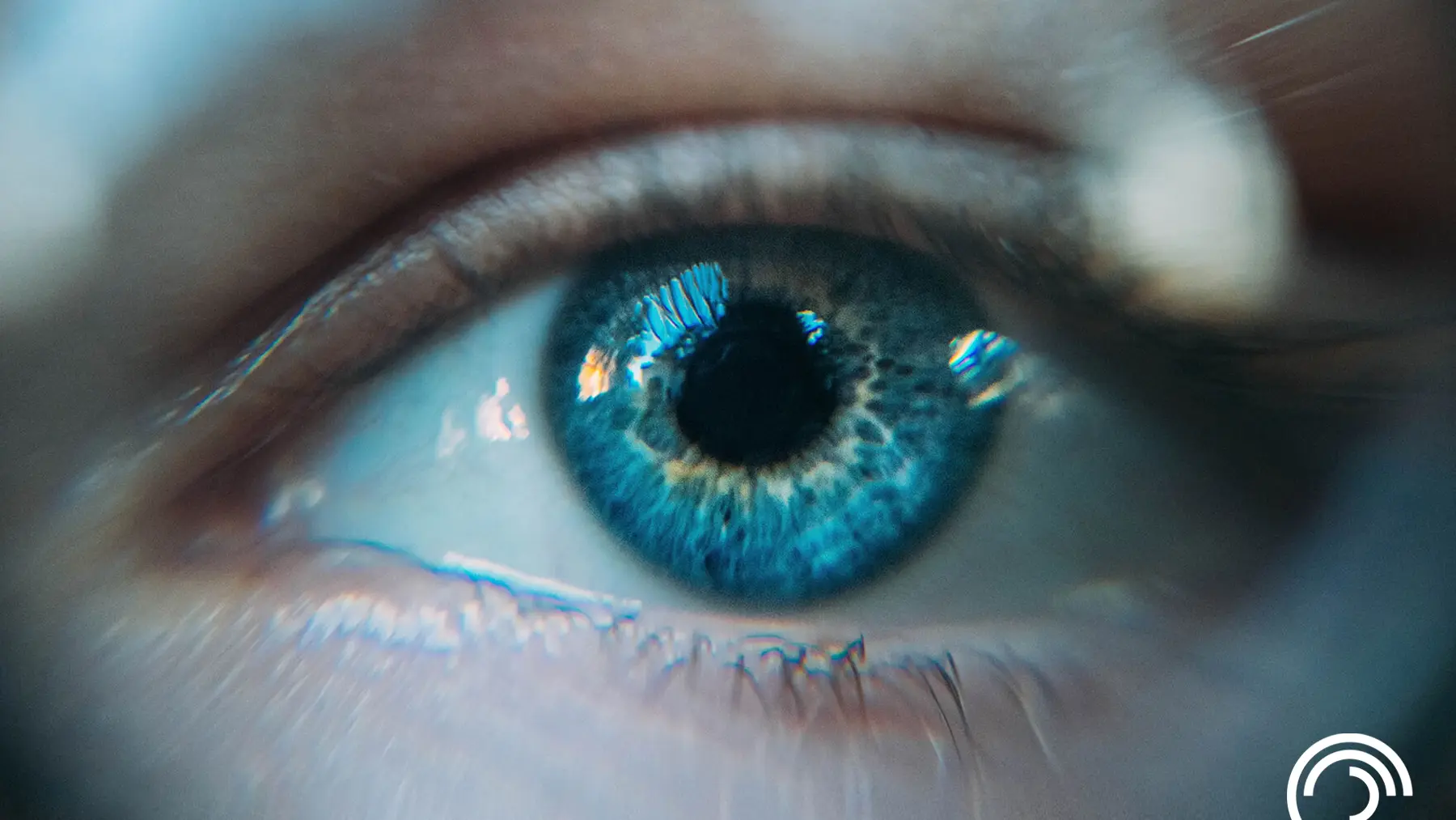 Close-up of a blue eye with intricate details, reflecting light. A logo is visible in the lower right corner.