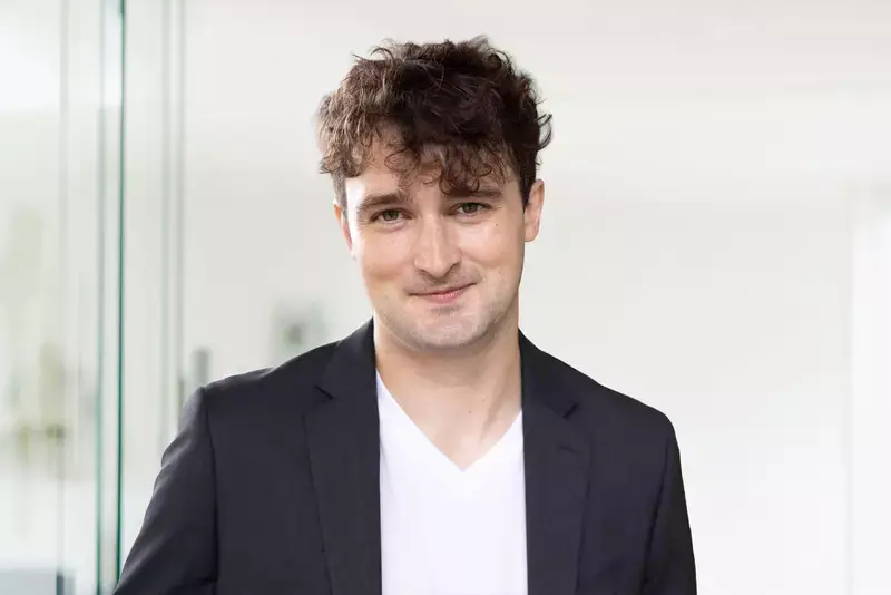 A person with curly hair wearing a black blazer and white shirt smiles slightly, standing in a bright room with glass in the background.