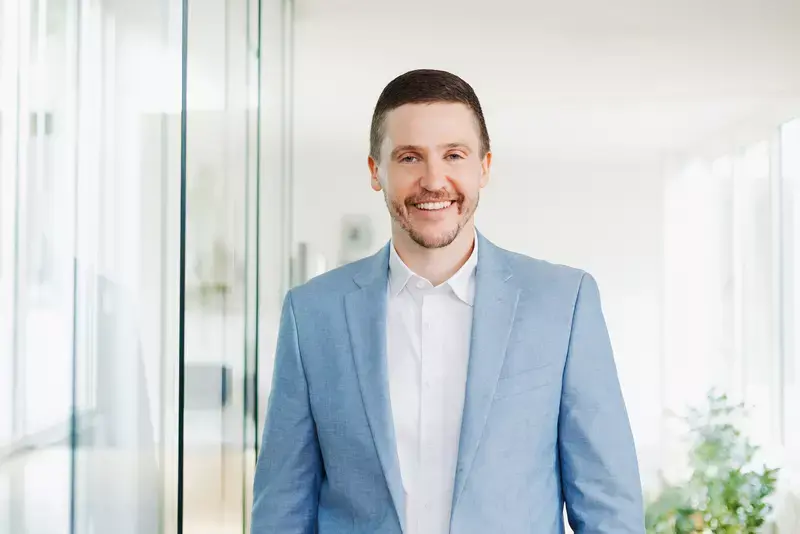 Man in a light blue suit and white shirt smiling in a bright, modern office with glass partitions and natural light.