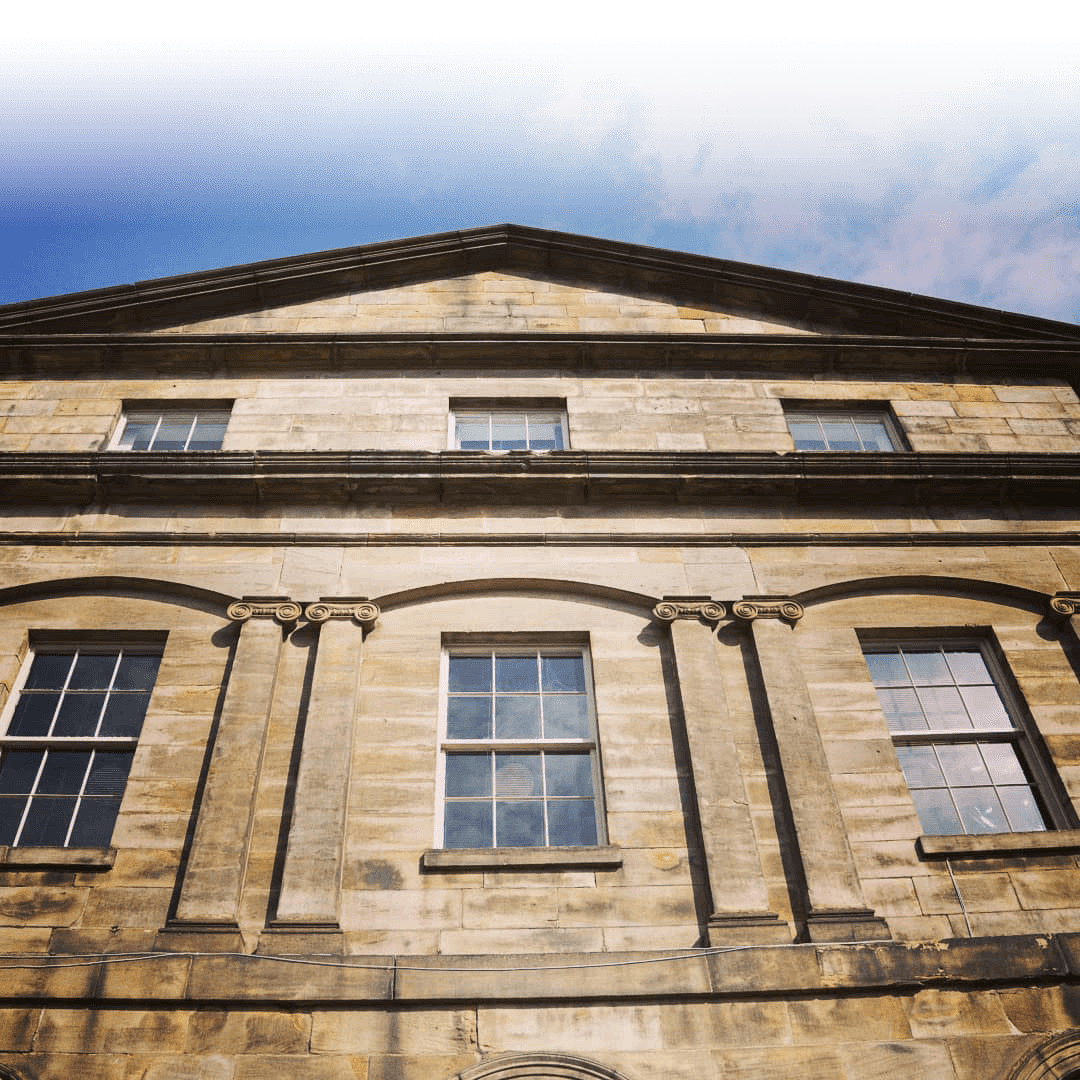 View of a classical stone building facade with tall rectangular windows and architectural detailing against a blue sky.