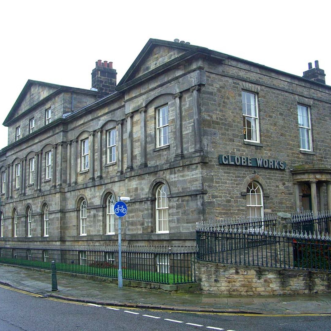 Historic stone building labeled "Globe Works," with arched windows and a small fence, situated on a corner street.