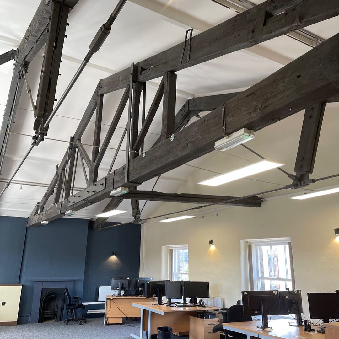 Open office with exposed wooden beams, modern desks with computers, soft lighting, and carpeted flooring.