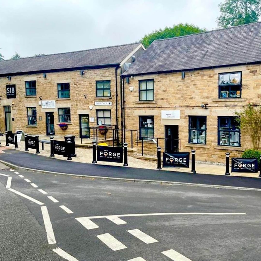 Stone buildings with shops line a street; black signs display "FORGE." The scene includes a paved walkway and a road with markings in the foreground.