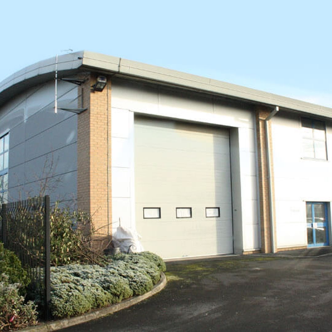 Modern commercial building with a large garage door, adjacent parking area, and landscaped greenery under a clear blue sky.