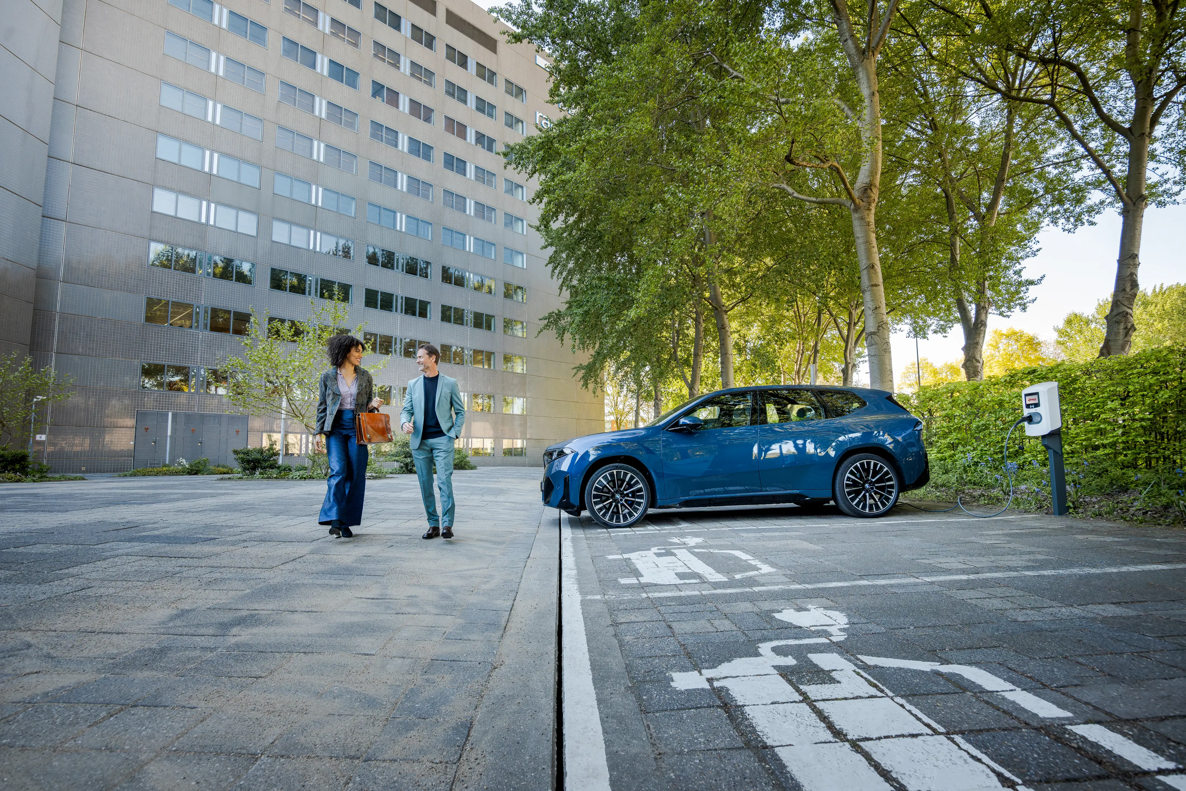 Two people walking near a parked blue electric car in a charging spot, with a modern high-rise and trees in the background.