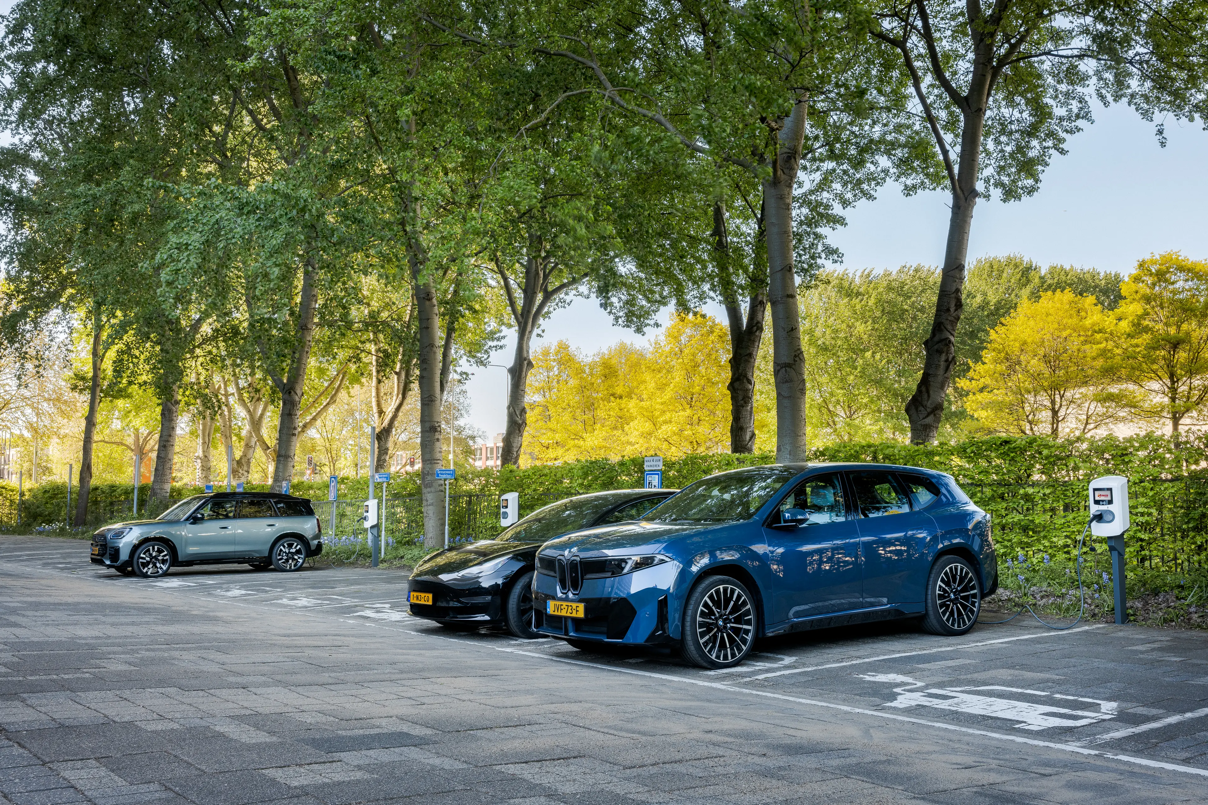 Electric cars parked and charging in a tree-lined parking area, with green foliage in the background.