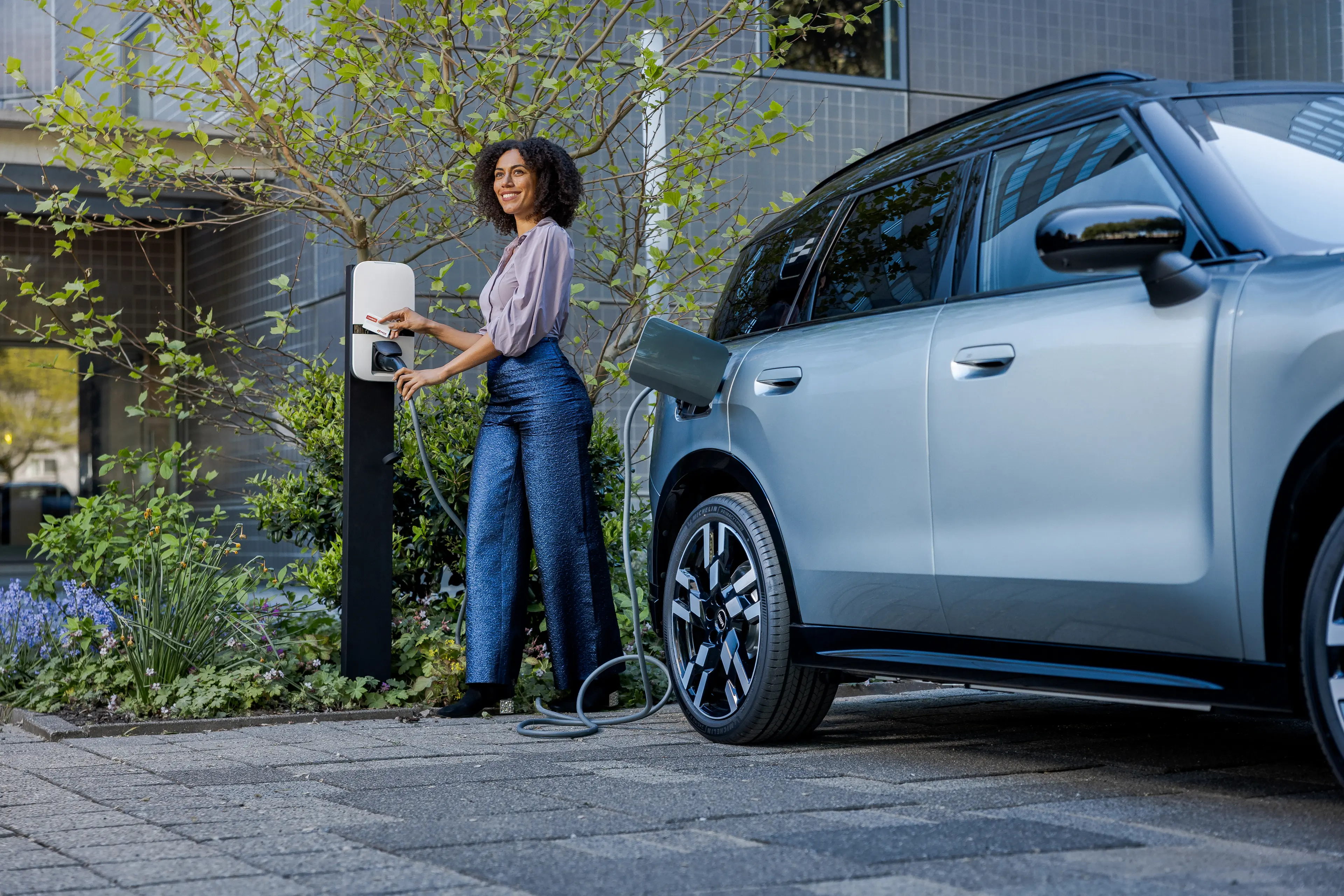 A woman charges an electric car at a charging station next to a modern building with greenery around.