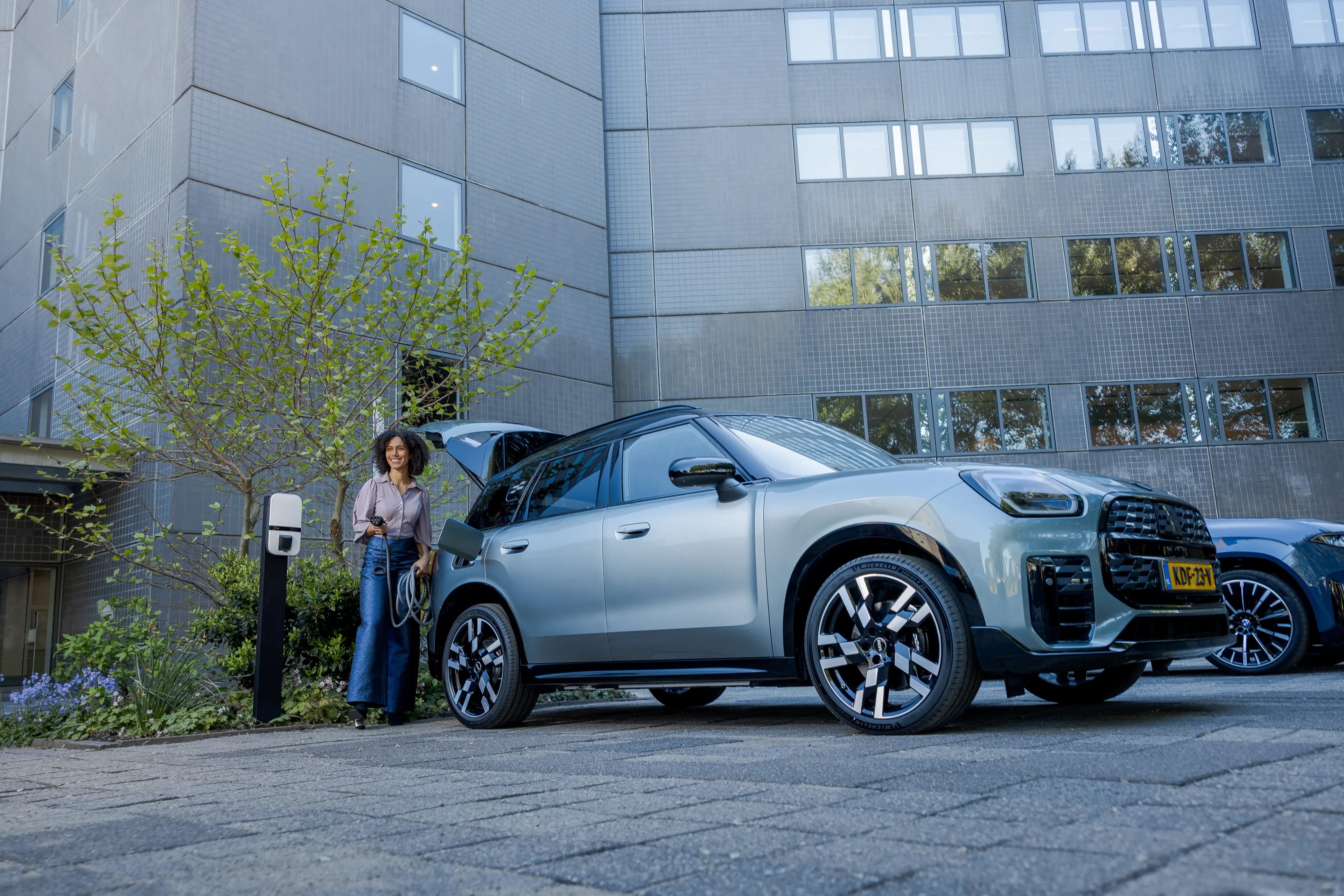 Woman standing by a silver SUV parked near a charging station in front of a modern building with reflective windows.