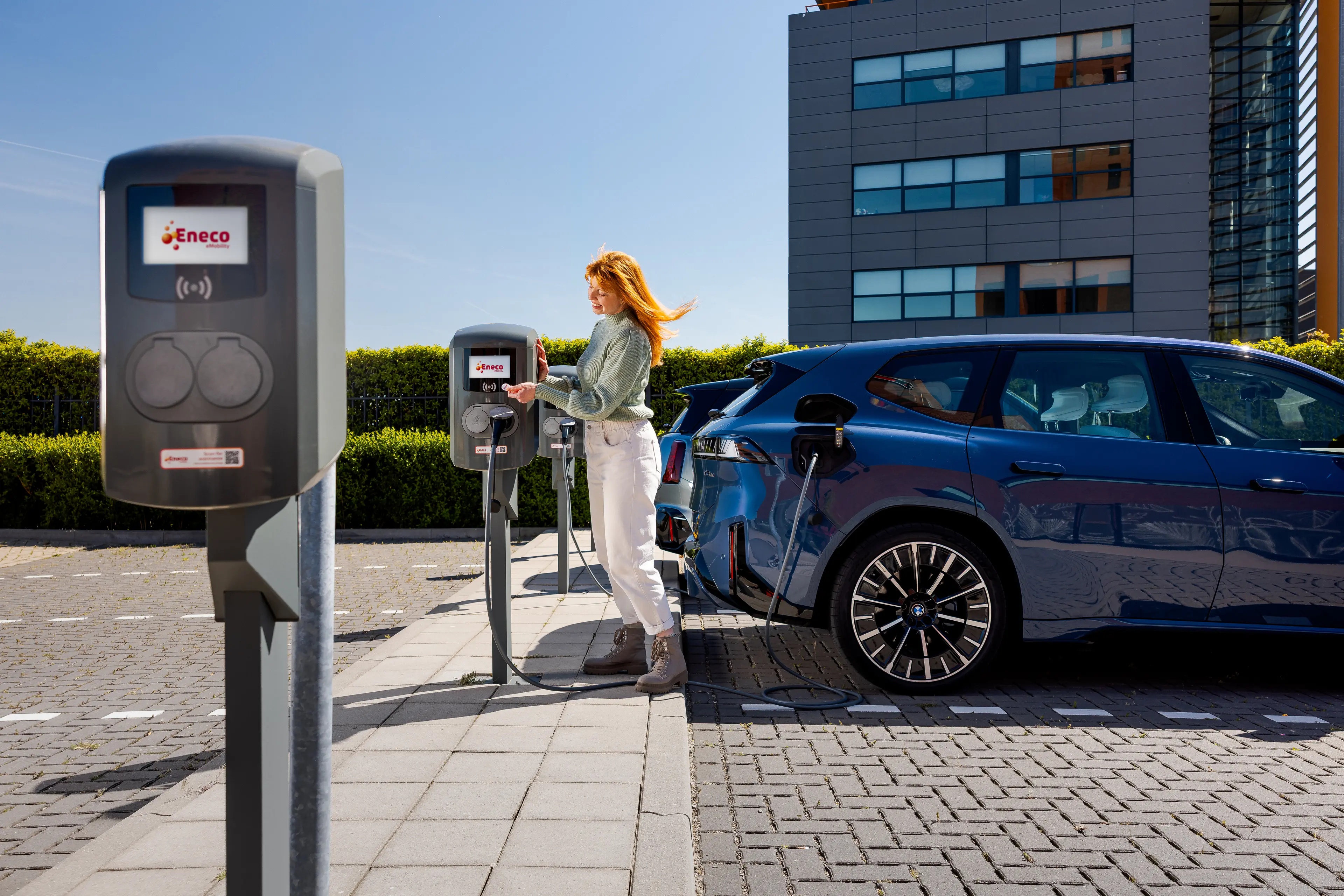 Person charging an electric car at a charging station in a parking lot, with a modern building in the background.