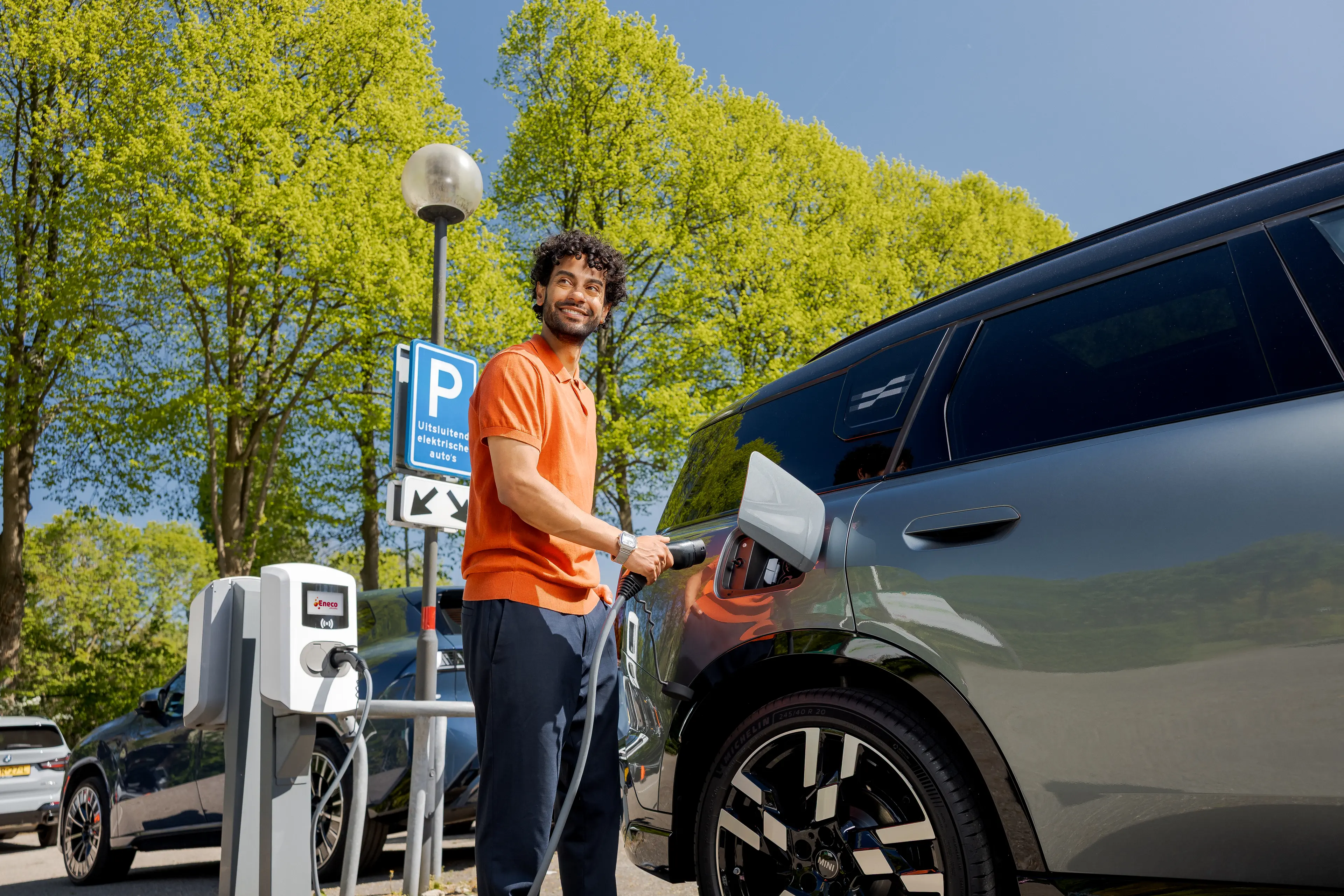 Man charging an electric car at a charging station in a sunny parking area with green trees in the background.
