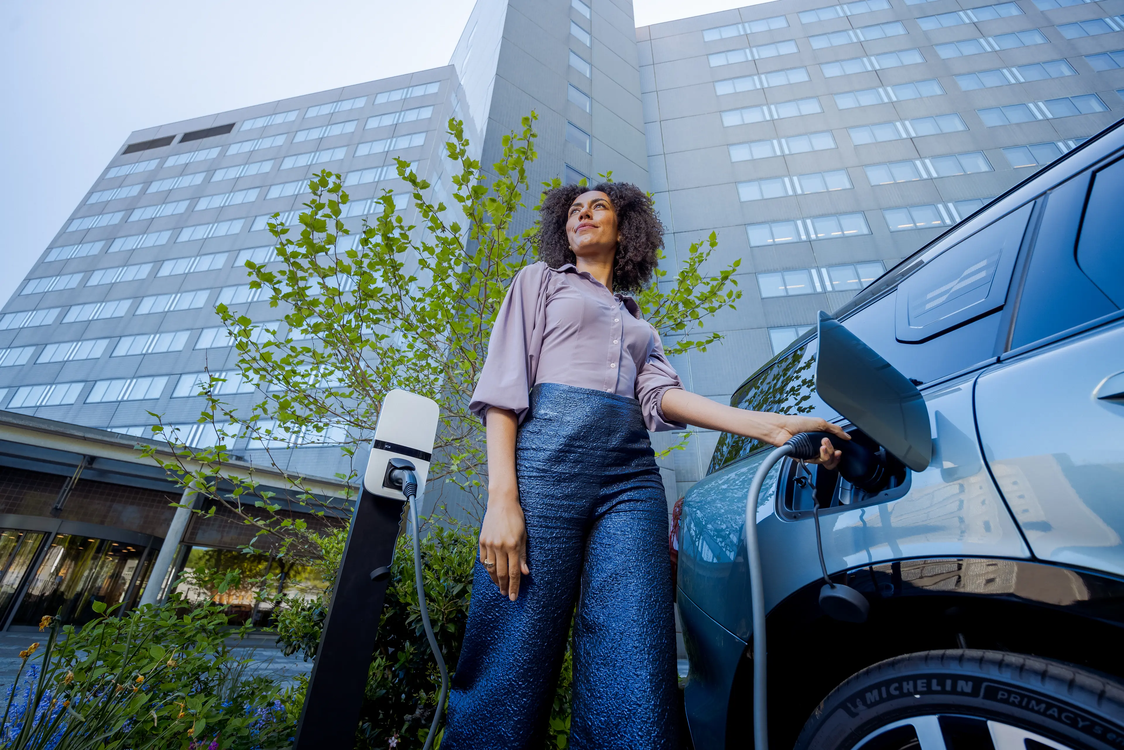 Woman standing by an electric car at a charging station in front of a modern building, looking upward.
