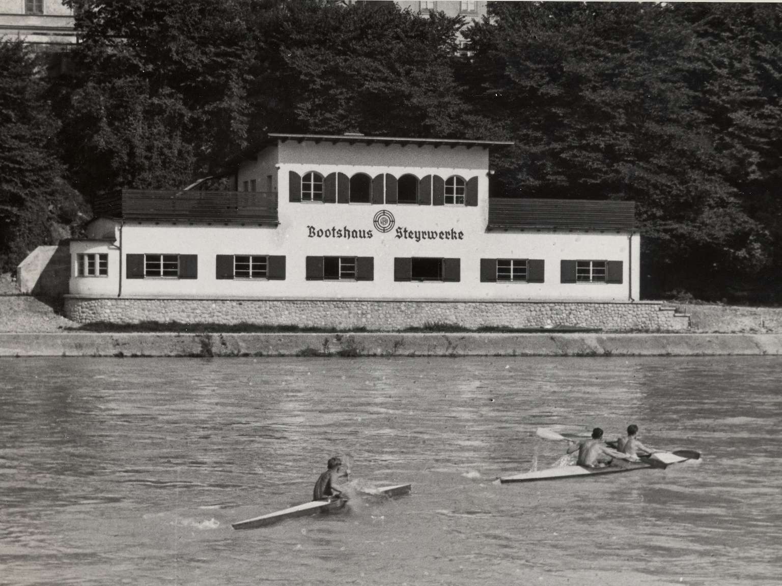 Schwarzweiß Foto des Bootshaus Steyrwerke am Ufer der Enns in der zwei Paddelboote mit Personen im Wasser treiben.