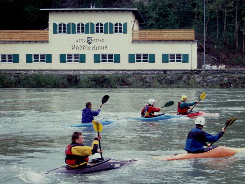 Abbildung des Paddlerhaus ATSV Steyr am Ufer der Enns mit mehreren Personen in Kajaks im Wasser.