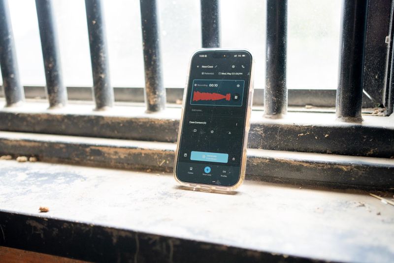 Smartphone leaning against a metal railing, with the CoVet mobile app recording a consultation, displaying a red waveform on its screen, possibly recording audio. Dusty windowsill in the background.