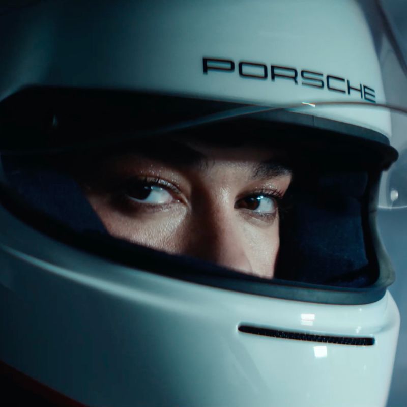 Dua Lipa schaut durch das offene Visier eines weißen Rennwagen-Helms mit der Aufschrift PORSCHE in die Kamera.