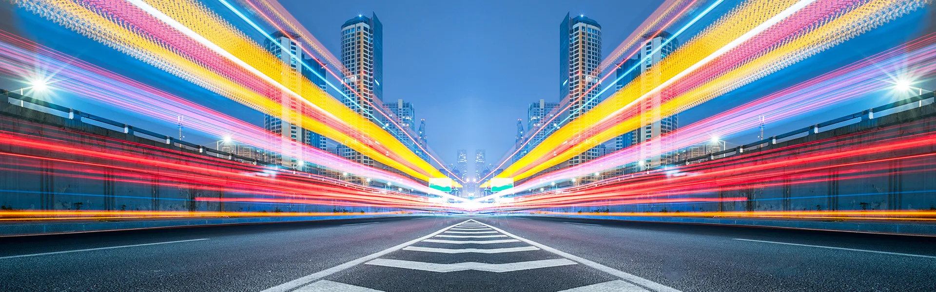 Urban night scene with colorful light trails from fast-moving vehicles, tall buildings, and a clear blue sky.