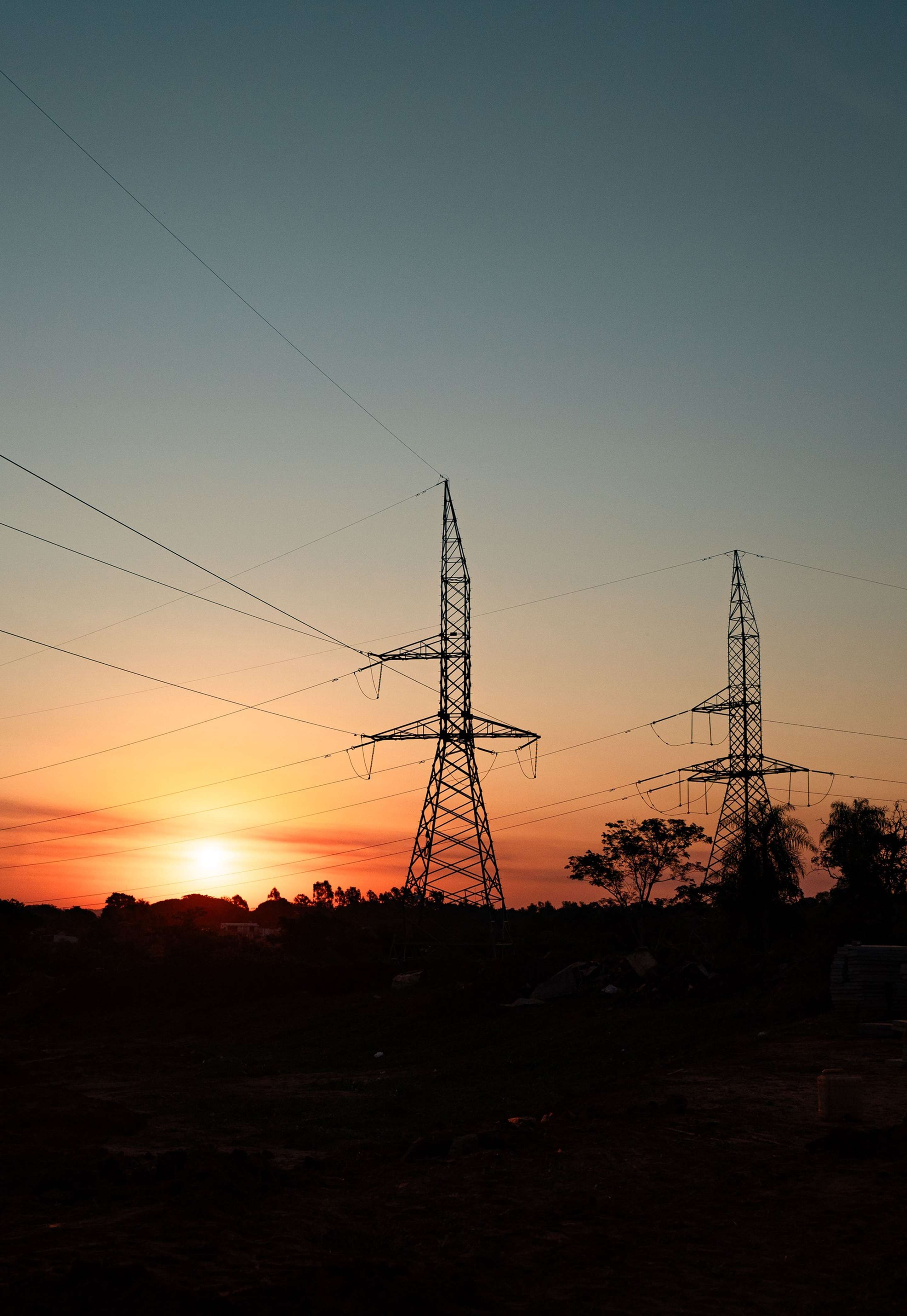 Silhouetted power lines and towers against a colorful sunset sky with scattered clouds and trees in the foreground.