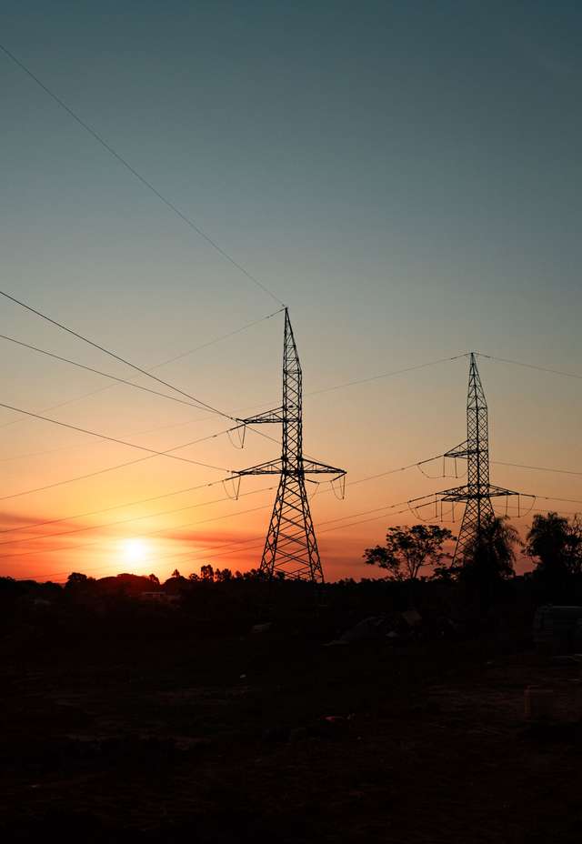 Silhouetted power lines and towers against a colorful sunset sky with scattered clouds and trees in the foreground.