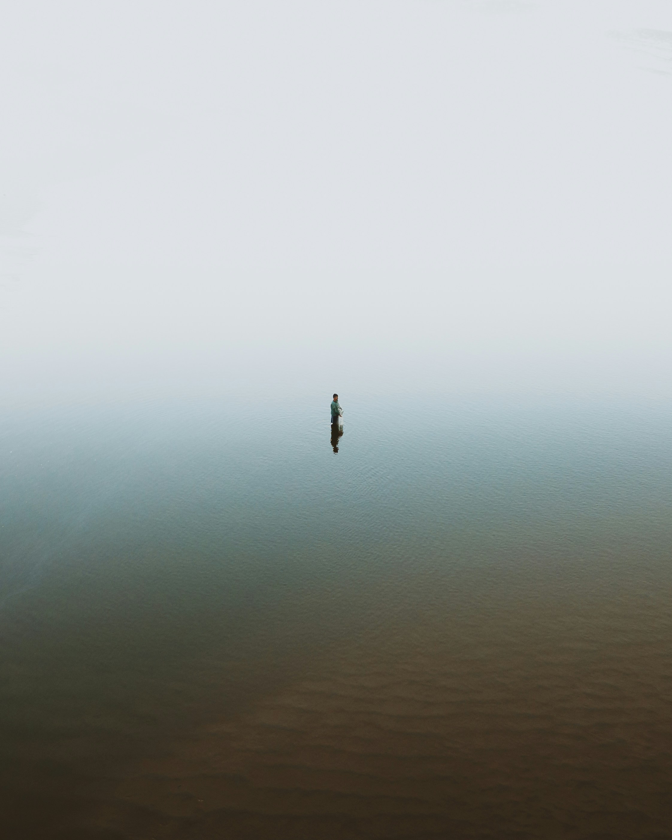 A lone fisherman stands waist-deep in a misty body of water during daytime, surrounded by fog, creating a serene and isolated atmosphere