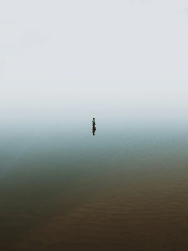 A lone fisherman stands waist-deep in a misty body of water during daytime, surrounded by fog, creating a serene and isolated atmosphere