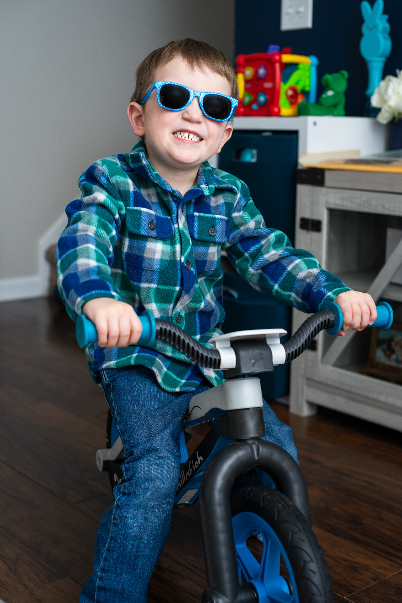 Smiling child wearing sunglasses and a plaid shirt rides a small bike indoors on a wooden floor.