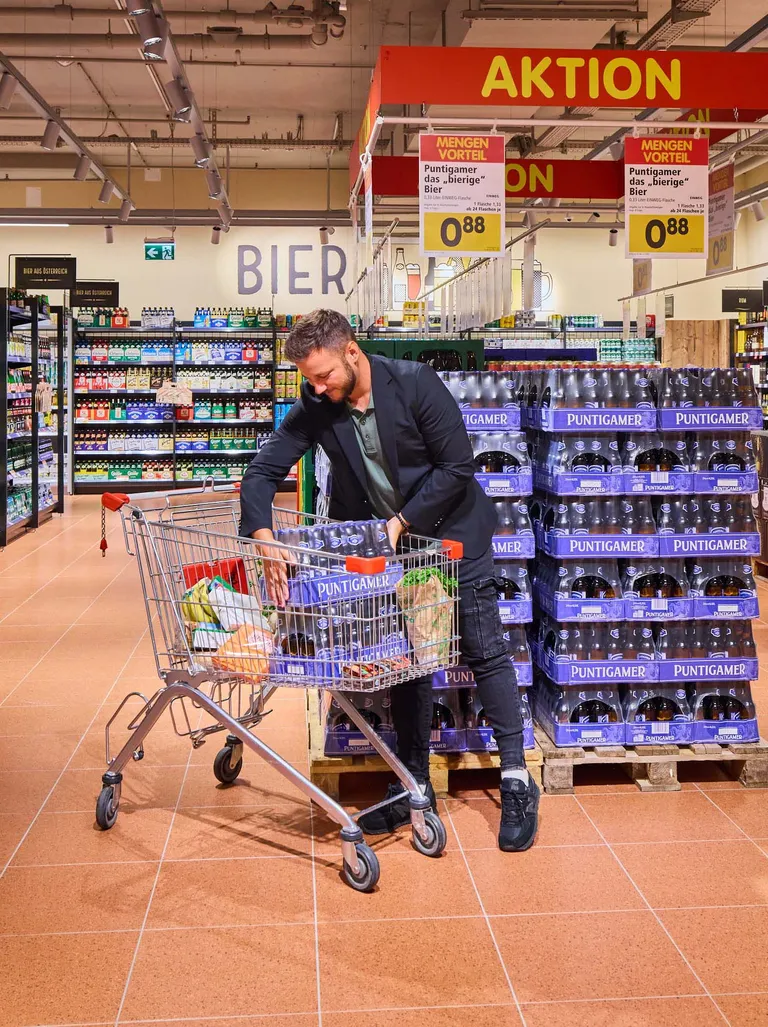 Ein Mann kauft in einem Supermarkt Bier ein und legt Puntigamer-Packungen in einen mit Lebensmitteln gefüllten Einkaufswagen. Schilder weisen auf ein Bier-Sonderangebot hin.