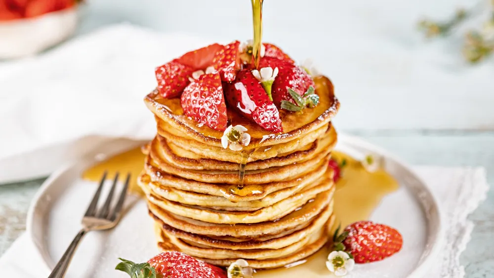 Ein Stapel Pancakes mit Erdbeeen und Blüten, auf die Ahornsitup gegossen wird