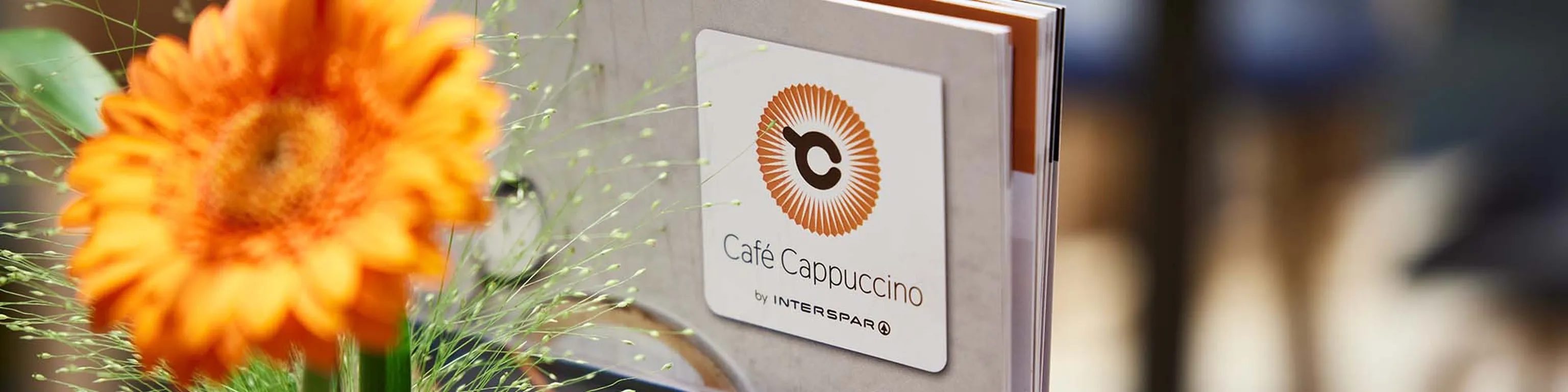 Nahaufnahme einer leuchtend orangefarbenen Blume neben einer Speisekarte des „Café Cappuccino“ mit Logo auf einer Holzfläche.