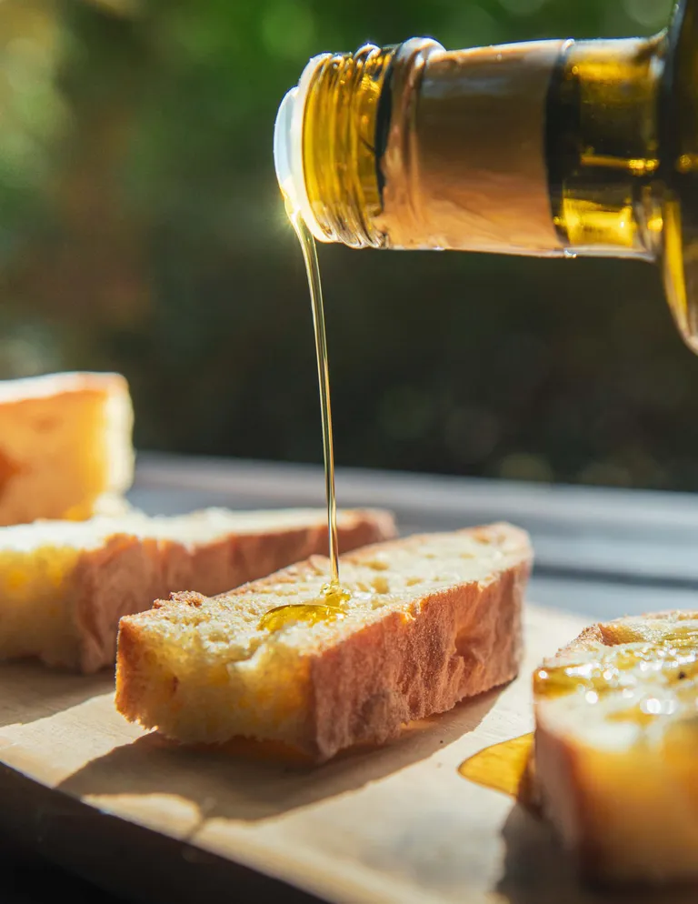 Auf einer Holzplatte wird Olivenöl über knusprige Brotscheiben gegossen, während Sonnenlicht die Szene erhellt.