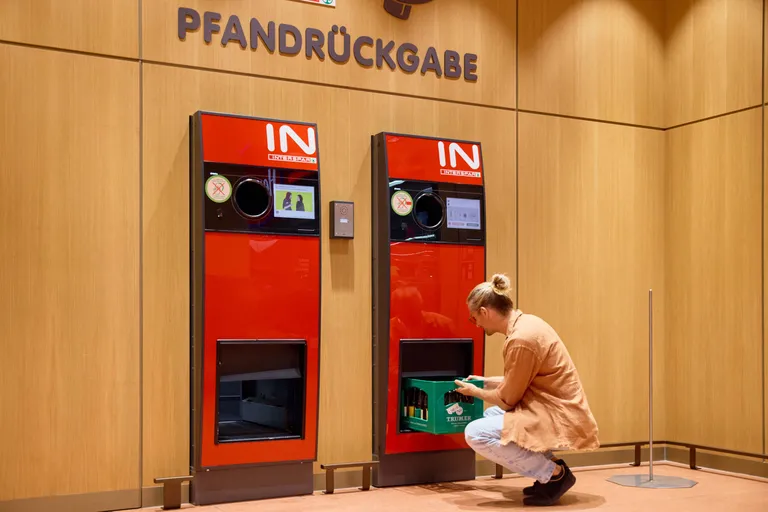 Eine Person, die Glasflaschen in einen roten Rücknahmeautomaten mit der Aufschrift „Pfandrückgabe“ in einer mit Holz vertäfelten Umgebung einwirft.
