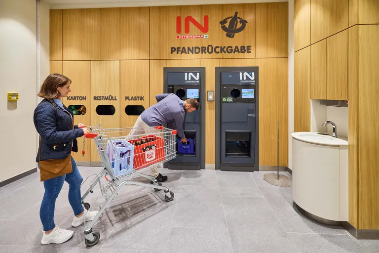 Eine Person bedient einen Flaschenrücknahmeautomaten bei INTERSPAR, während eine andere mit einem Einkaufswagen voller recycelbarer Flaschen wartet.