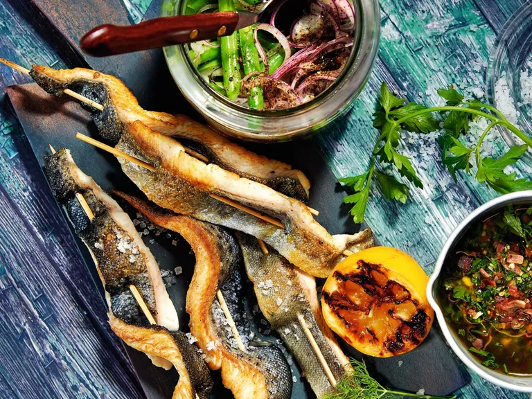 Gegrillter Steckerlfisch mit Speck-Pesto und Fisolensalat auf einem bläulichen Holztisch