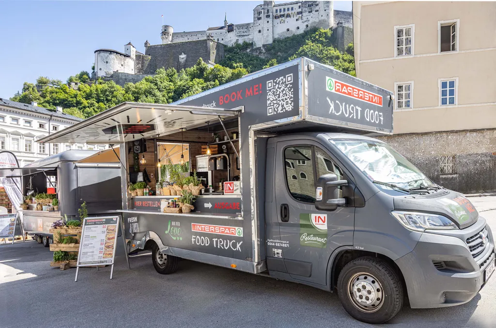 Ein grauer Imbisswagen mit dem „INTERSPAR“-Logo, der frische Produkte und Gerichte anbietet, steht in sonniger Lage in der Nähe einer Burg und eines beigen Gebäudes.