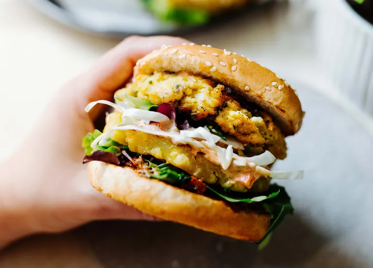 Eine Hand, die einen leckeren Wels-Burger mit Sesambrötchen, Blattgemüse, knusprigem Patty, Kohlstreifen und Soße hält.