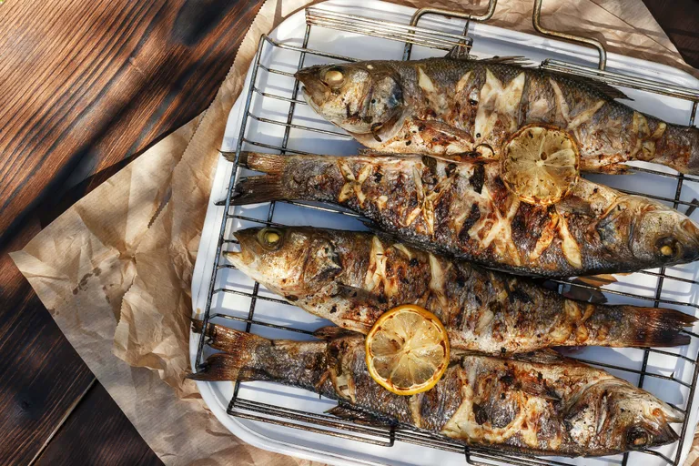Ein ganzer Fisch, auf einem Rost gegrillt, mit knuspriger Haut, garniert mit einer gerösteten Zitronenscheibe, im Sonnenlicht beleuchtet.
