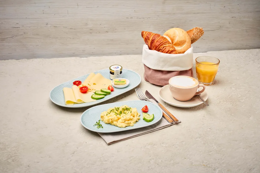 Frühstücksszene mit Rührei, Käsescheiben, Tomaten, Gurken, Croissants, Brot, Butter, Cappuccino und Orangensaft auf einem hellen Tisch.
