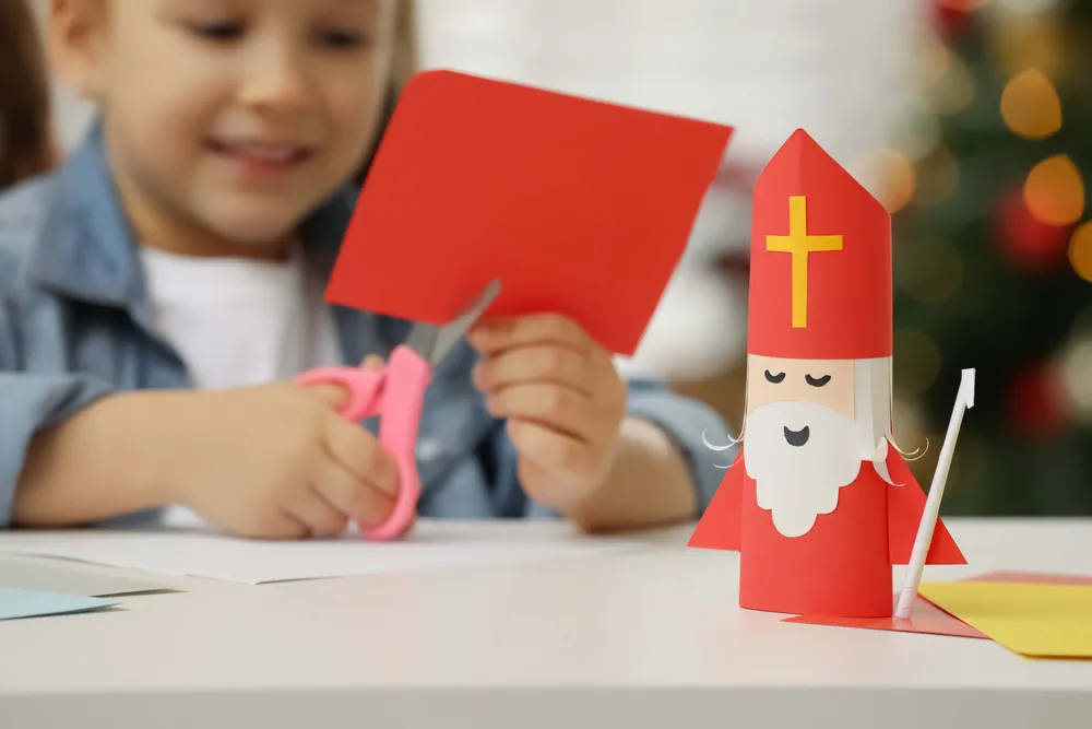 Child crafting with red paper, using scissors. A paper figure resembling Saint Nicholas is on the table.