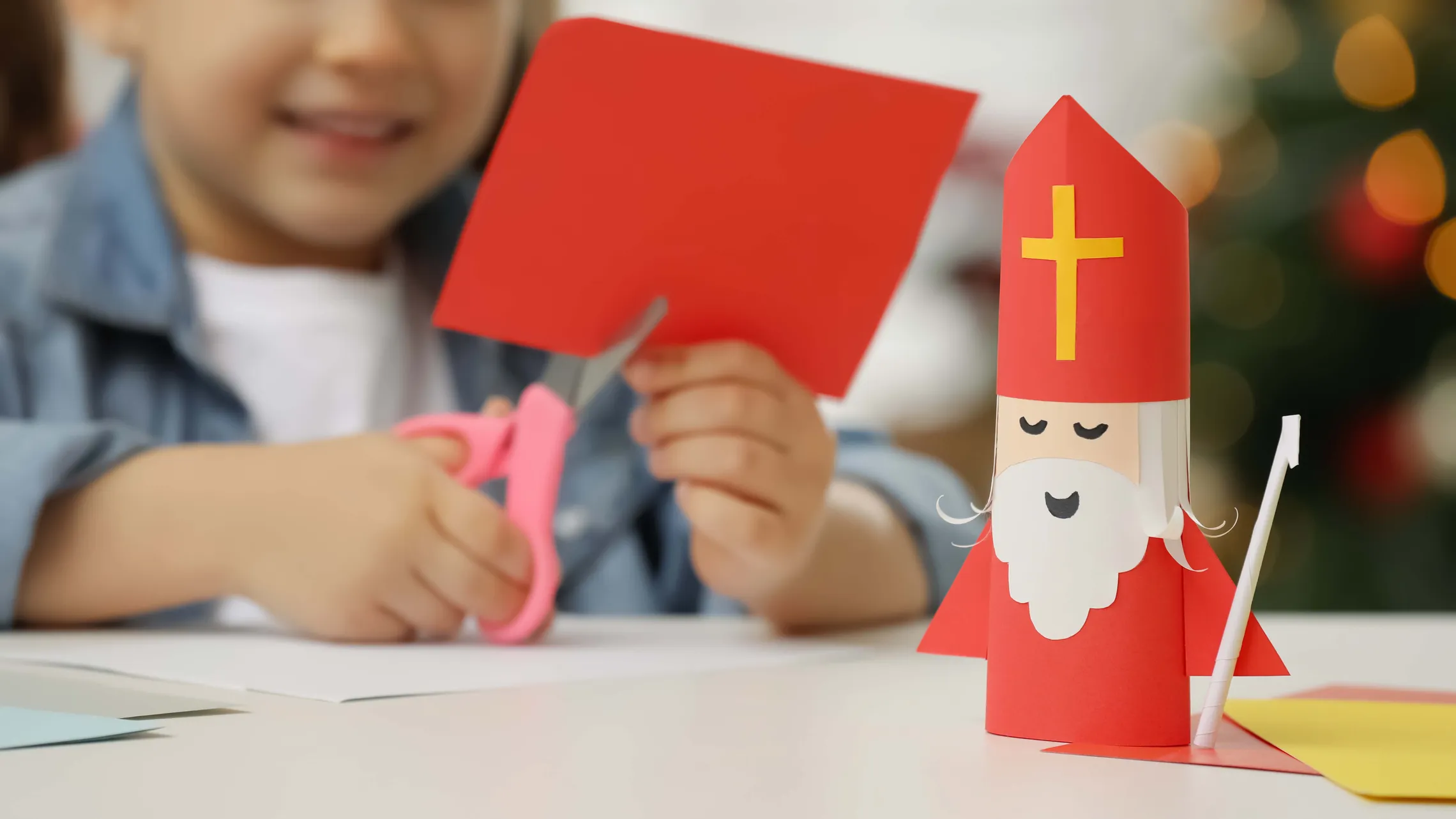 Child crafting with red paper, using scissors. A paper figure resembling Saint Nicholas is on the table.