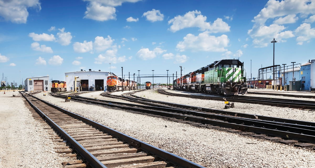 Rail yard with several tracks and points in the foreground, orange and green diesel locomotives parked in front of buildings under a blue sky.